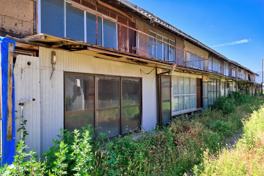 放置されて空き家問題の実態となっている状況をイメージした、手前に雑草が生い茂り、外壁やベランダに老朽化が見られる、青空の下に佇む古い木造の長屋（アパート・集合住宅）