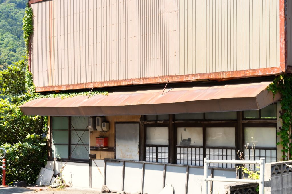 長期間放置されて空き家問題の実態となっている物件をイメージした、錆びついた大きなひさしや年季の入った引き戸があり、外壁につる植物が絡まり始めている、山間の緑を背景に佇む古い木造の空き家（元店舗・住宅）