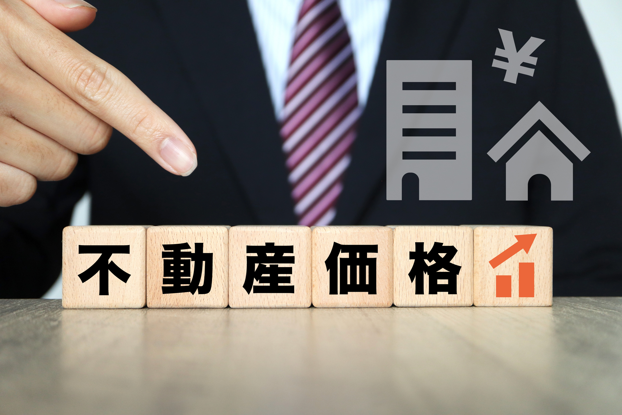 横一列に並ぶ『不動産価格』と上昇グラフが描かれた木製ブロック、それを指し示すビジネスパーソンの指、ビルや住宅、円マーク（￥）のイラスト