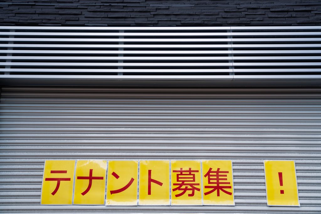 使われていない遊休不動産（店舗やオフィスなど）を賃貸に出して有効活用することをイメージした、閉められた金属製のシャッターに、黄色い紙に赤文字で『テナント募集！』と一文字ずつ書かれた貼り紙