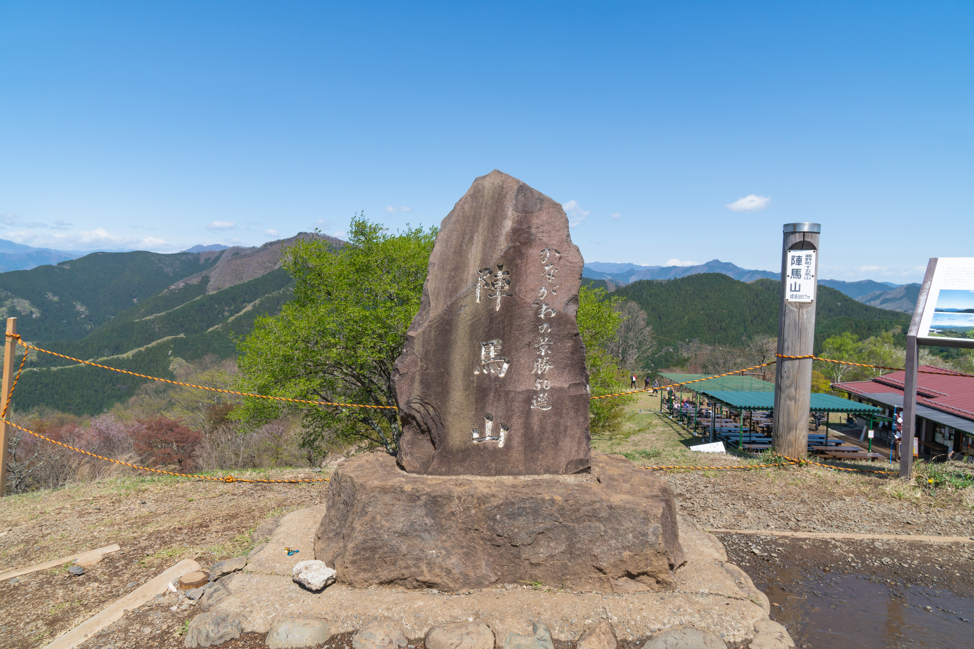 青空の下、陣馬山の山頂に佇む「陣馬山」と「かながわの景勝50選」の文字が刻まれた大きな石碑。背景には緑豊かな山々の連なりと、右側に木製の標識や茶屋の建物が写っている風景写真。