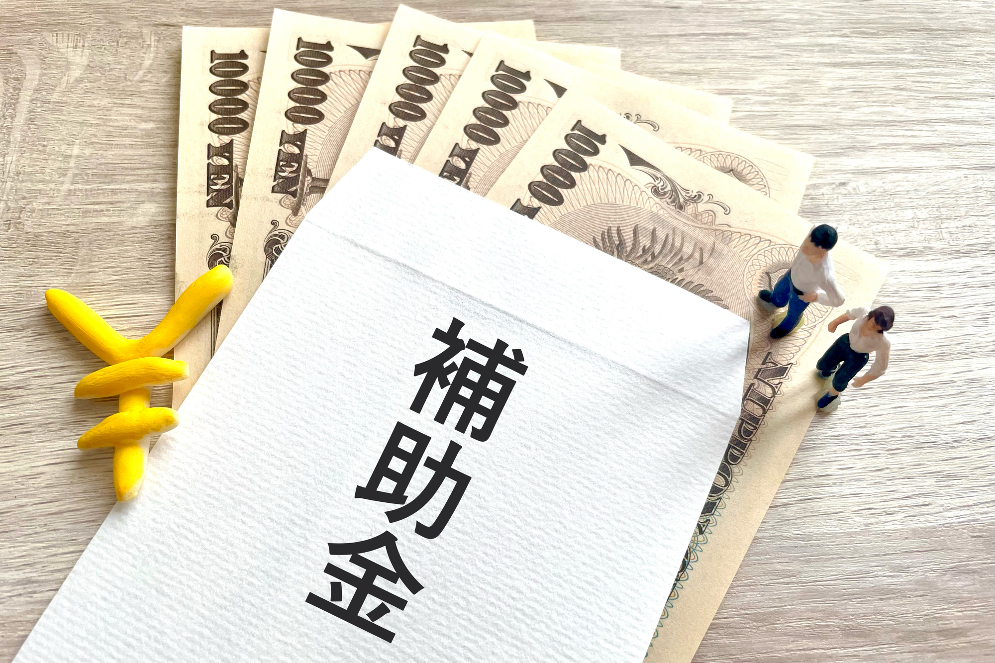 「補助金」と書かれた封筒と一万円札、人のミニチュア