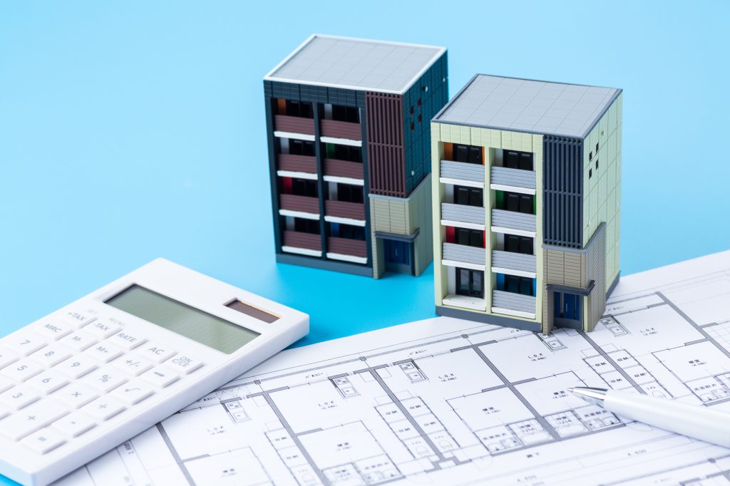 マンションの間取り図の上に置かれた2棟のマンション模型と電卓。マンションの規模や専有面積に応じた修繕積立金の相場計算をイメージした写真。