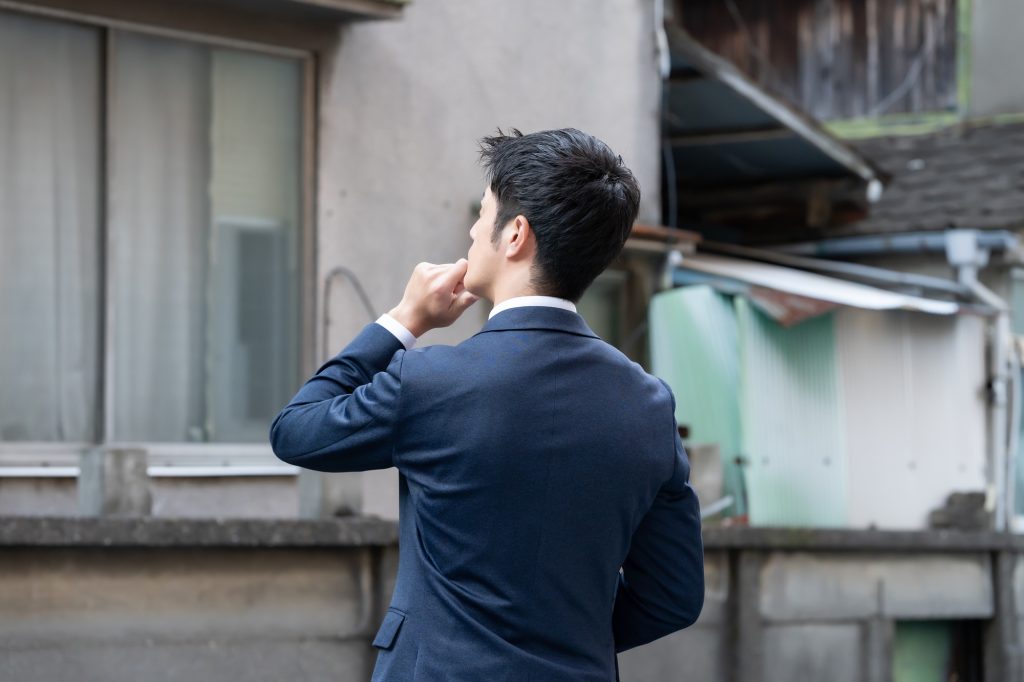 中古住宅購入時の注意点や内見時の確認ポイントをイメージさせる、古い住宅の前で思案するスーツ姿の男性の後ろ姿