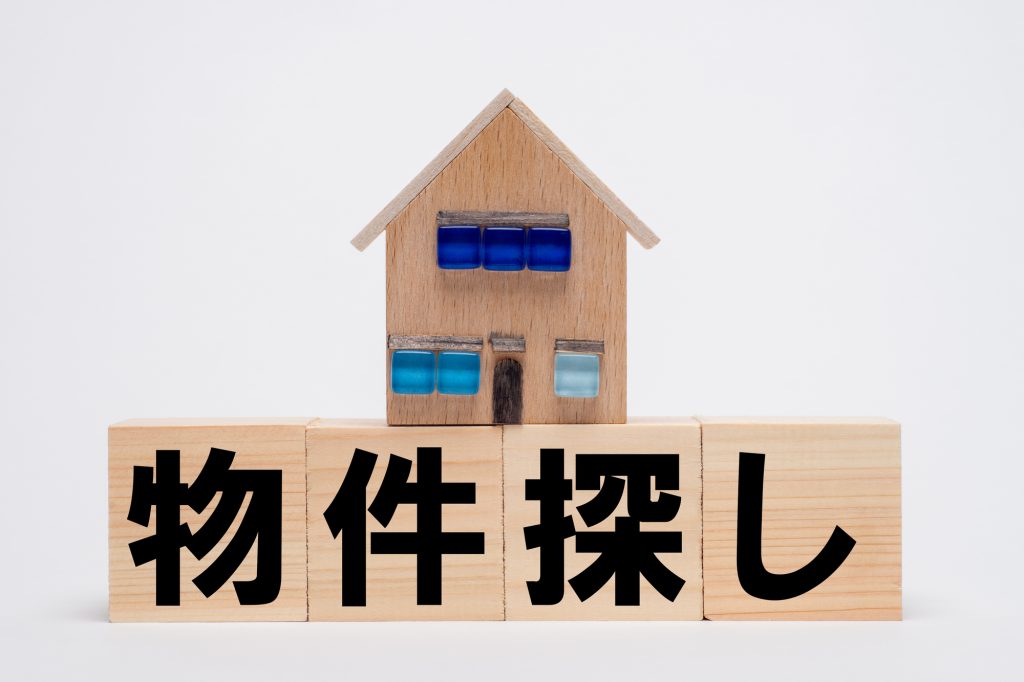「物件探し」の文字が書かれた木製ブロックの上に載る家型オブジェ(倉庫の売り物件探しやリサーチのイメージ)