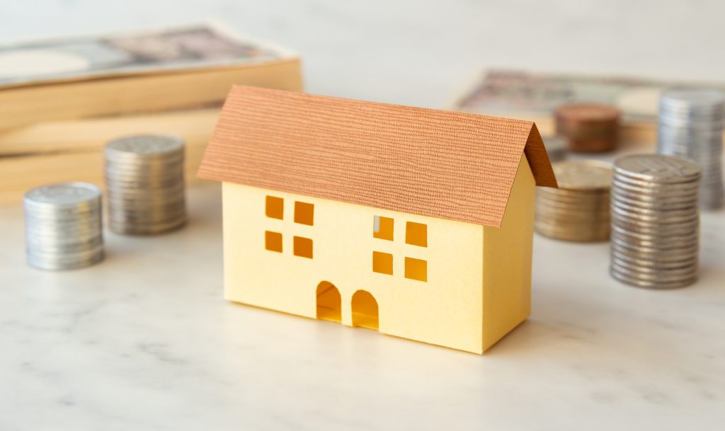 アパート投資の資金計画をイメージした、積み上げられた硬貨や紙幣の束の前に立つ住宅模型の画像