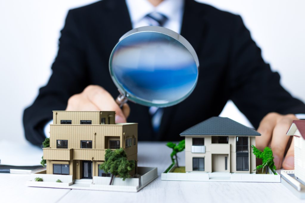 複数の住宅やマンションのミニチュア模型を虫眼鏡でのぞき込むビジネスパーソン（不動産投資における利回り相場や物件選定のイメージ）