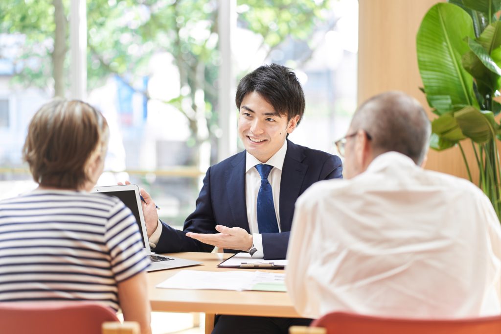 タブレットを使いながら笑顔で説明するスーツ姿の男性と相談する夫婦(倉庫購入時の融資の受け方と必要書類のイメージ)