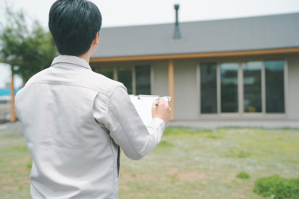 クリップボードを持ち一戸建て住宅を見つめる作業着姿の男性(現地調査や建物点検のイメージ)
