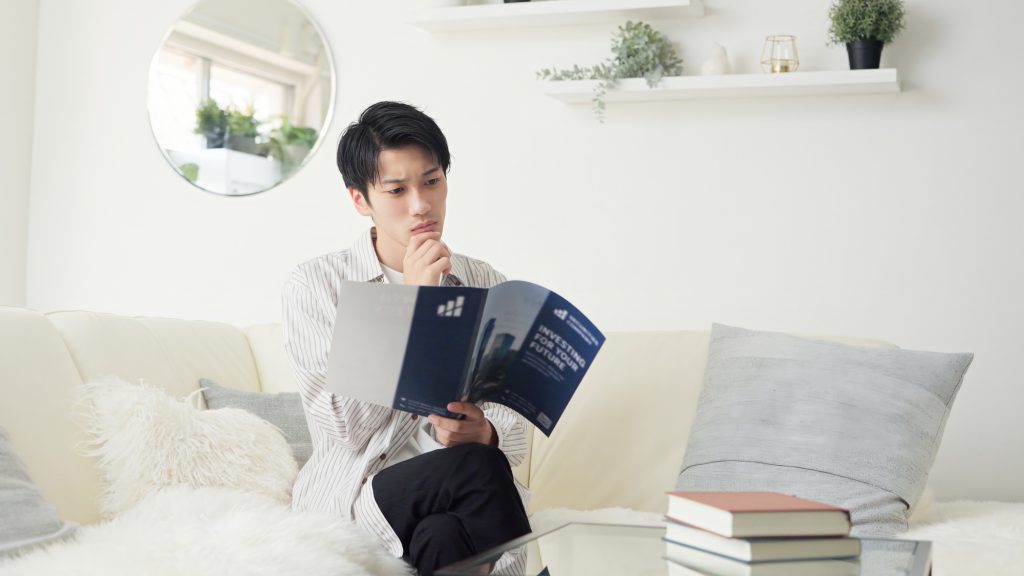ソファに座り、悩むような表情で資料を読み込む男性（不動産選びや運用の検討のイメージ）