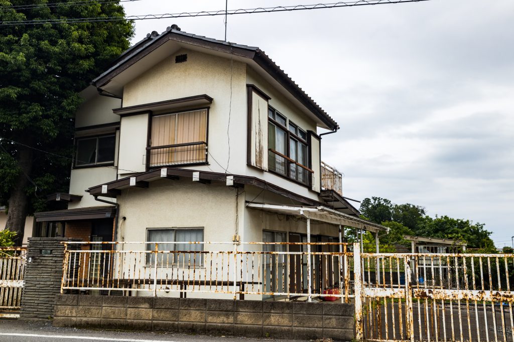 錆びた白いフェンスとブロック塀に囲まれた、築年数の経過した2階建ての木造一軒家